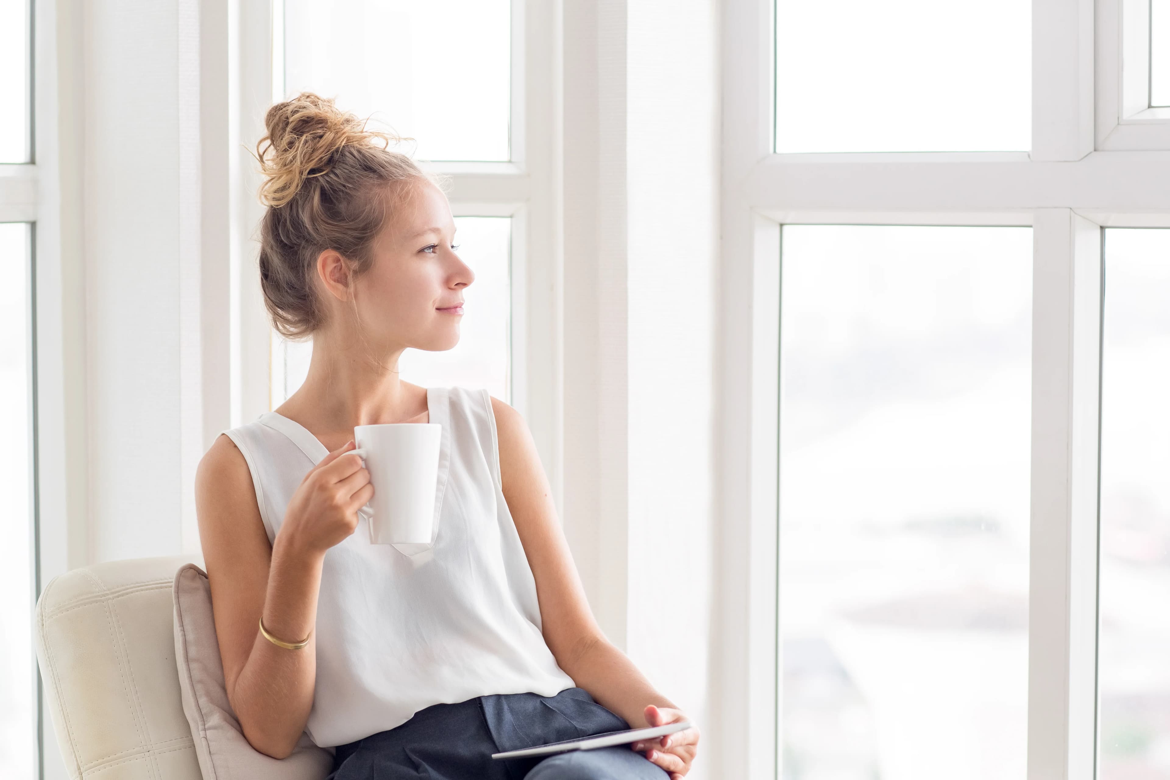 Femme détendue, une tasse à la main, devant la fenêtre de son salon